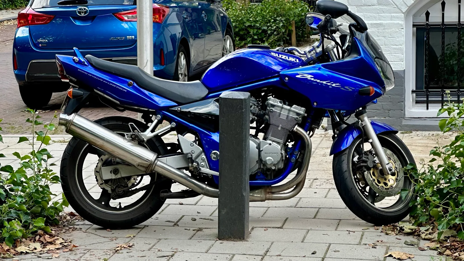 A blue motorcycle with a large silver exhaust parked beside cars on a leafy residential street in Utrecht.