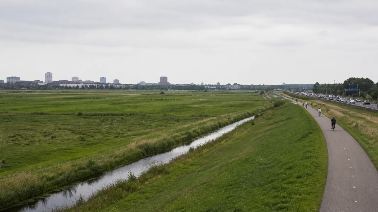 Green fields and a drainage canal in Rijnenburg beside a cycle path and a busy road, with Utrecht's urban edge in the distance.